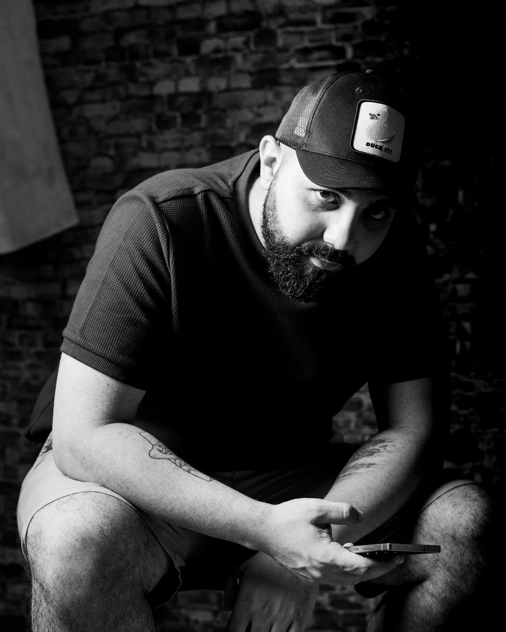 Ritratto ambientato in bianco e nero: uomo seduto con cappello contro un muro di mattoni